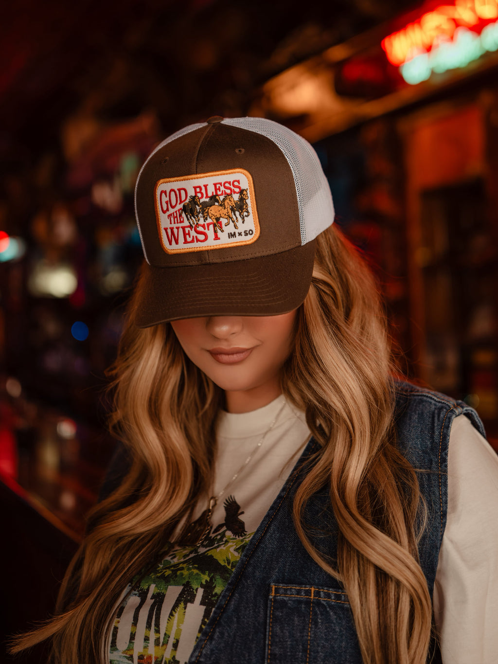 Brown trucker cap with 'God Bless the West' patch, worn by person with long hair