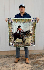 Wild rag scarf held up outdoors, featuring cowboy on horse with colorful floral edge.